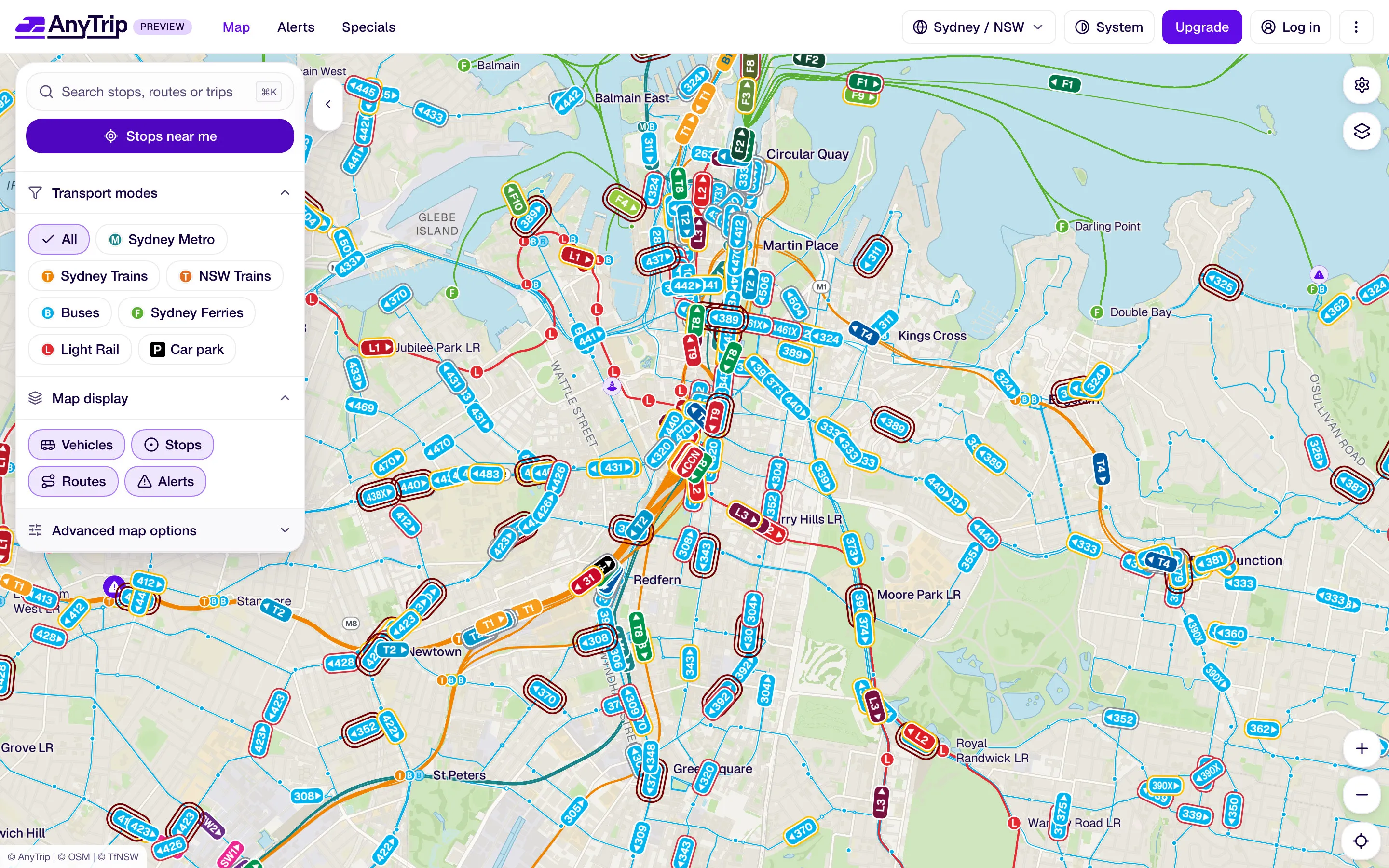 The AnyTrip web app’s live map of inner Sydney, with train, metro, bus, ferry and light rail vehicles shown as route badges along their lines. A side panel offers stop search, a “Stops near me” button, transport mode filters and map display options for vehicles, stops, routes and alerts.