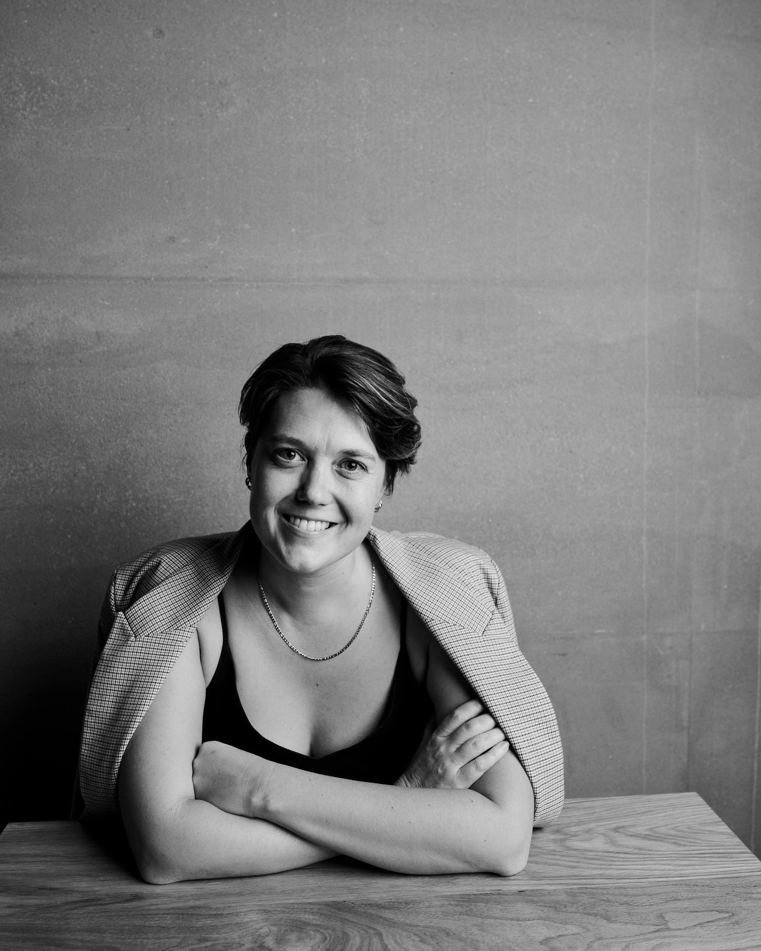 Black-and-white portrait of Stella Blake-Kelly, smiling, seated with her arms folded on a wooden table